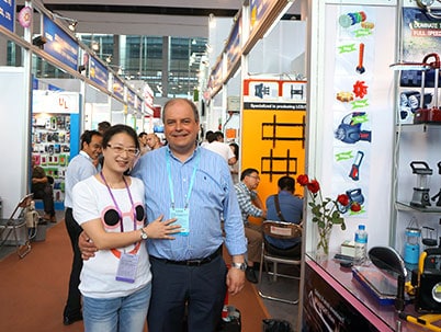 Two people smile together in an indoor market with booths and other attendees visible in the background.