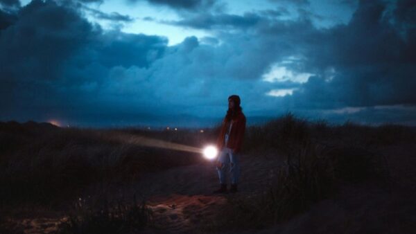 A person with a flashlight stands among grassy dunes at dusk under a cloudy sky.