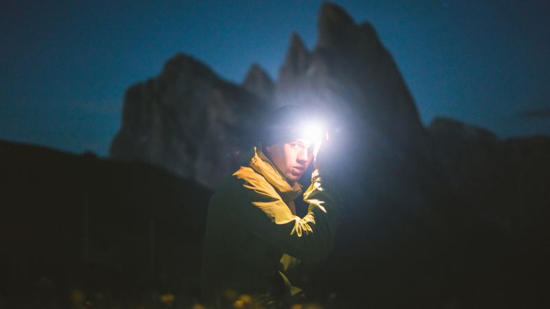 Une personne portant une veste jaune et une lampe frontale se tient dehors au crépuscule, avec des montagnes floues en arrière-plan.