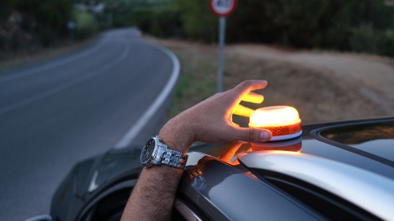 Bright orange emergency warning light on top of a vehicle during daytime roadside scene with a person's hand operating the light, emphasizing road safety and alert vehicle features.